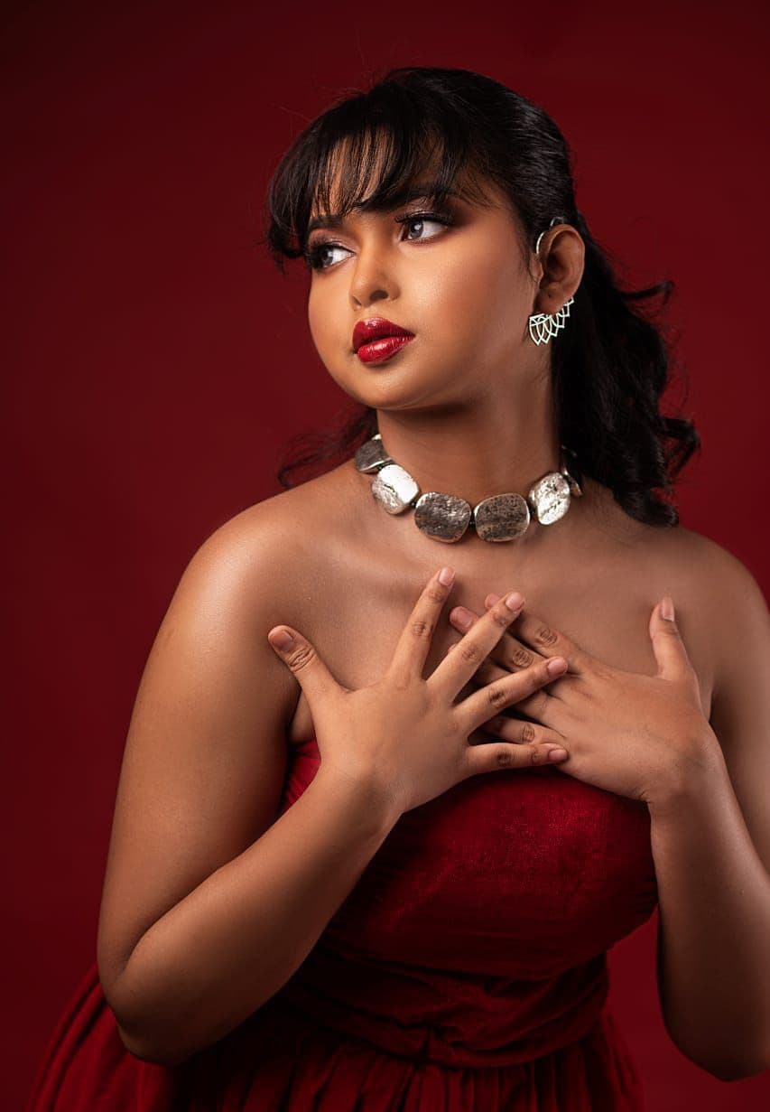 Sanjana Murmu in a red strapless gown with a silver plate choker, looking off camera against a deep red studio ground.