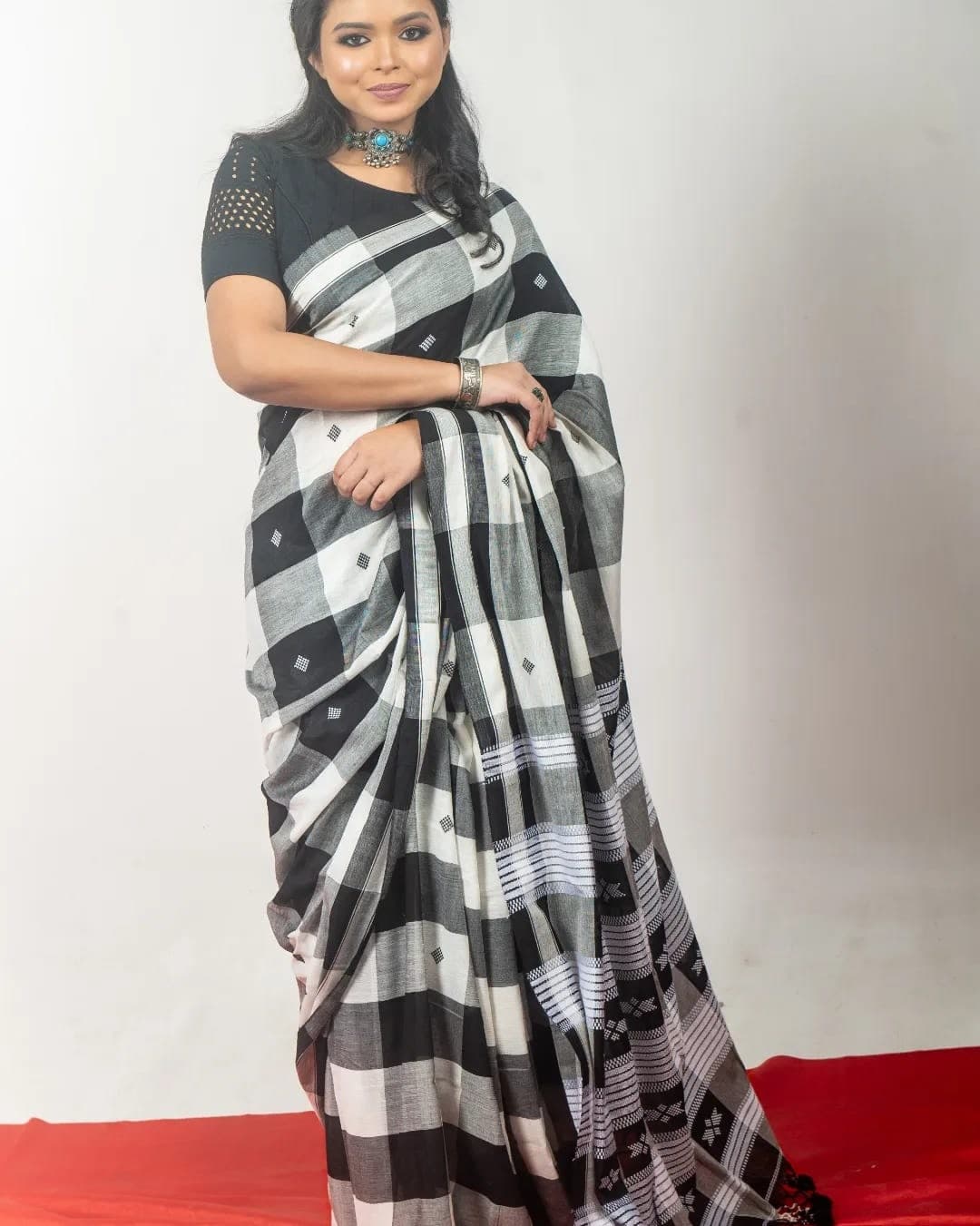 Sanjana Murmu in a black and white checked silk saree with a black blouse, one hand at her waist, against a pale studio ground.