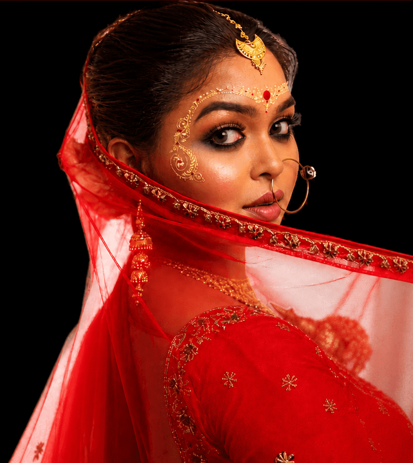 Sanjana Murmu looking over her shoulder beneath a red gold-bordered dupatta, with gold forehead ornament and nose ring.