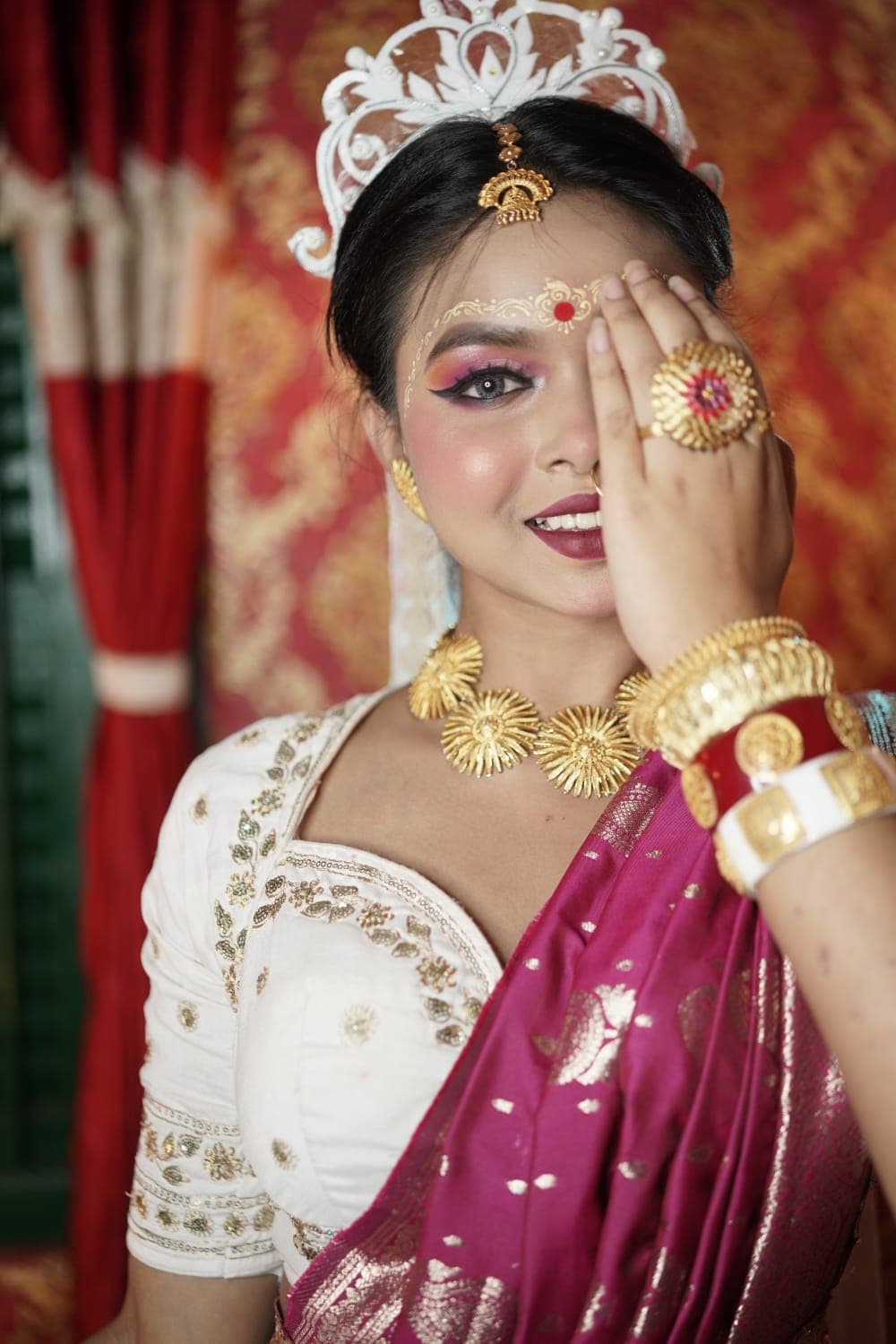 Sanjana Murmu holding a gold flower ring over one eye, smiling, in a magenta and white festive saree.