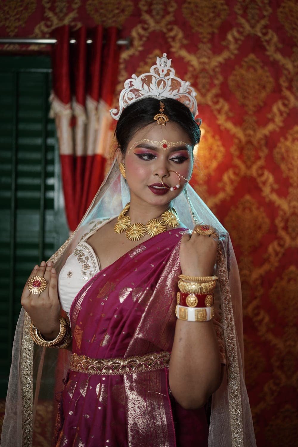 Sanjana Murmu with a hand raised to her shoulder, veil behind, in magenta and gold festive dress.