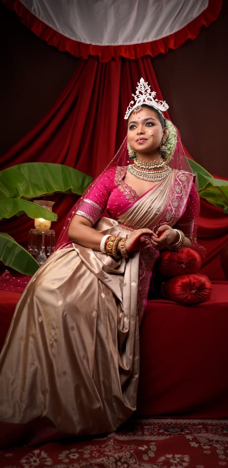Sanjana Murmu seated in a beige and gold saree with a white crown, red drapes and brass behind her.
