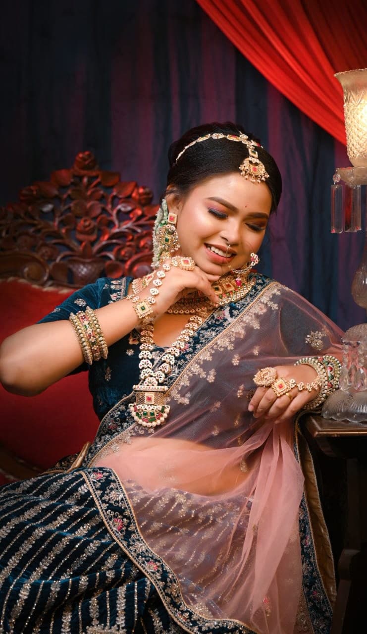 Sanjana Murmu seated against carved wood in a navy velvet lehenga and blush dupatta, hand raised to her chin, a lantern lit beside her.