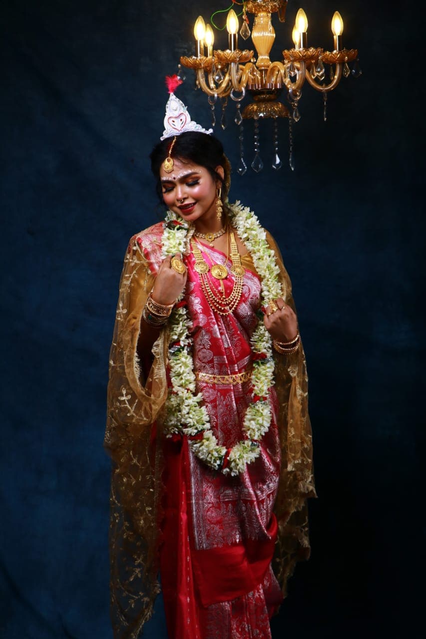 Sanjana Murmu in a red and gold Benarasi saree with a white topor and jasmine garland, standing beneath a lit chandelier against a deep teal ground.