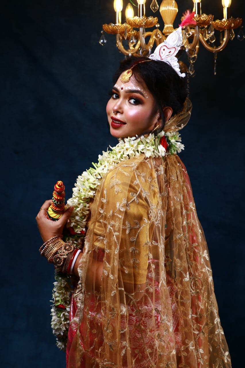 Sanjana Murmu in a red and gold Benarasi saree with a white topor and jasmine garland, holding a ritual ornament, gold dupatta falling from her shoulder.