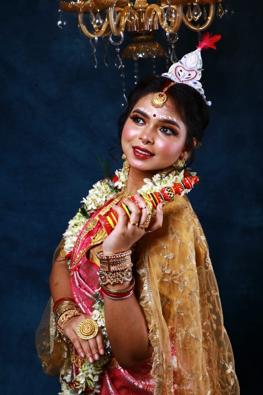 Sanjana Murmu in a red and gold Benarasi saree with a white topor and jasmine garland, one hand raised, looking directly ahead beneath a chandelier.