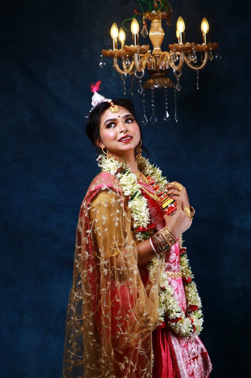 Sanjana Murmu in a red and gold Benarasi saree with a white topor and jasmine garland, turned slightly away, sheer gold dupatta draped across her arm.