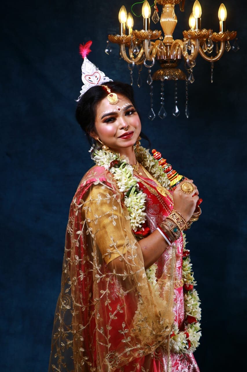Sanjana Murmu in a red and gold Benarasi saree with a white topor and jasmine garland, arms folded, gold dupatta drawn around her shoulders.
