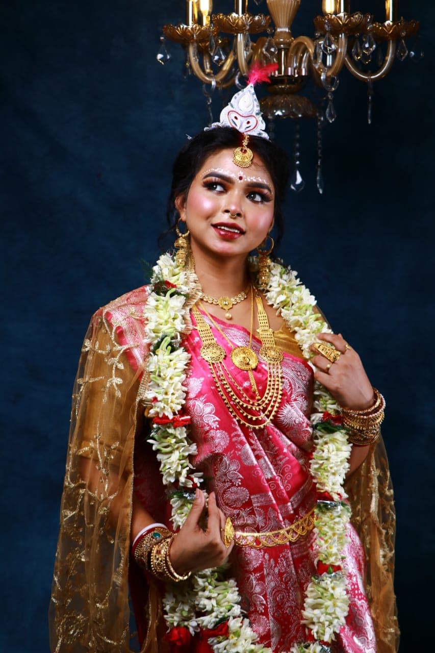 Sanjana Murmu in a red and gold Benarasi saree with a white topor and jasmine garland, standing centred, garland and layered gold necklaces at her chest.