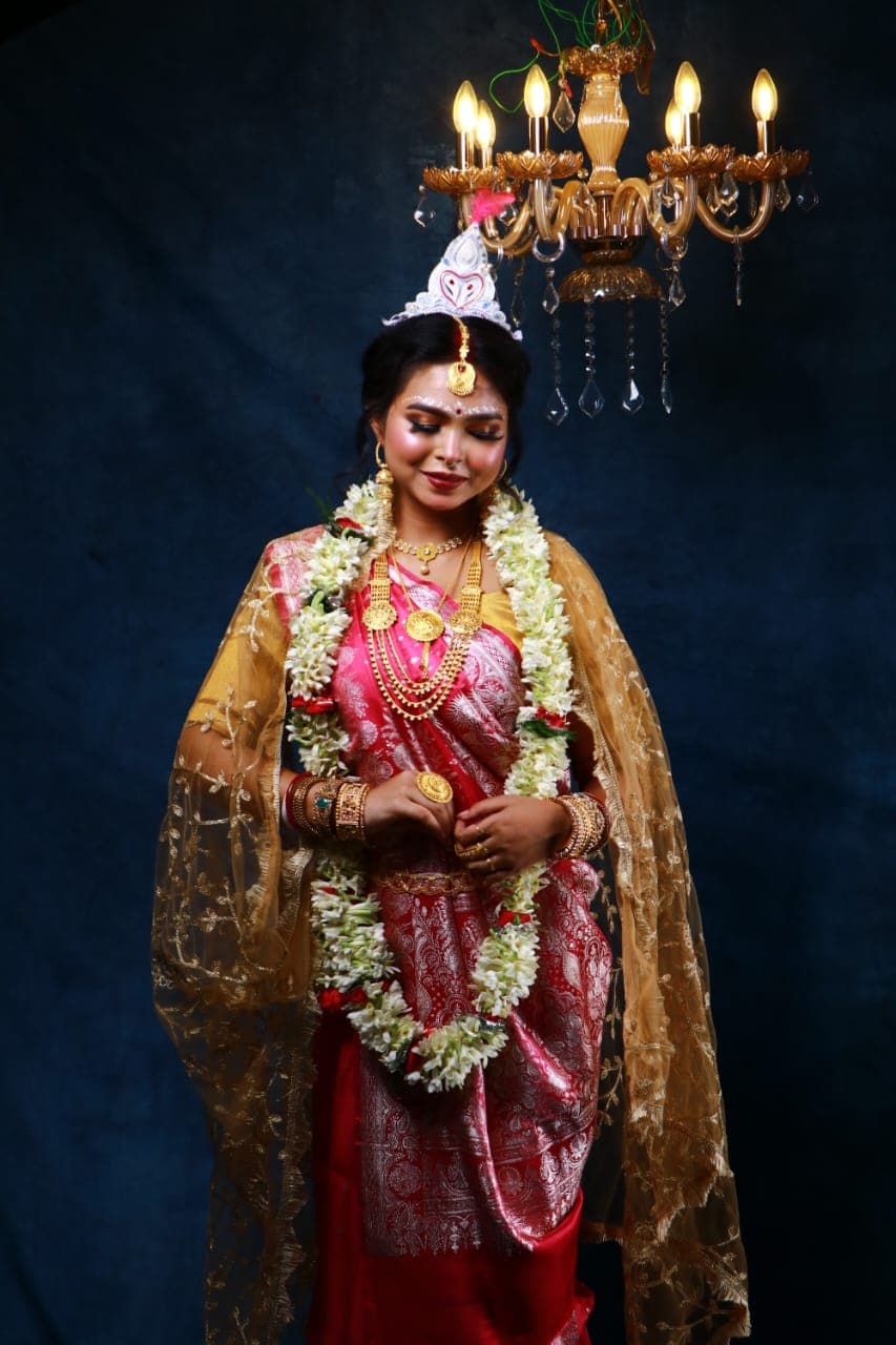 Sanjana Murmu in a red and gold Benarasi saree with a white topor and jasmine garland, holding the edge of her dupatta, chandelier lit above.
