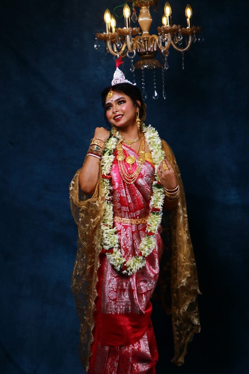 Sanjana Murmu in a red and gold Benarasi saree with a white topor and jasmine garland, hand raised to her shoulder, jasmine garland and gold bangles.