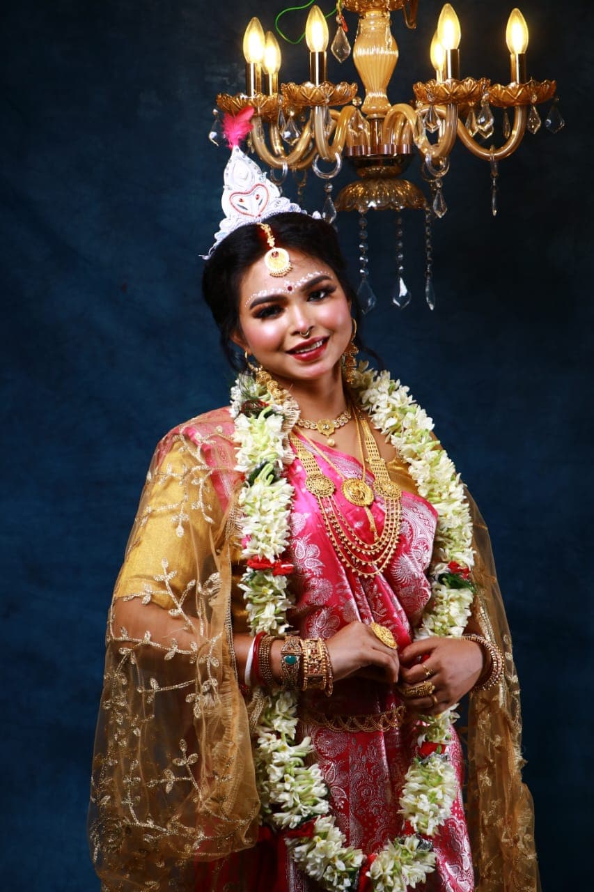 Sanjana Murmu in a red and gold Benarasi saree with a white topor and jasmine garland, hands together at her waist, gold dupatta falling behind.